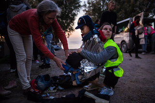 En nyanländ pojke fryser och värms med en filt medan volontärer söker passande kläder åt honom.